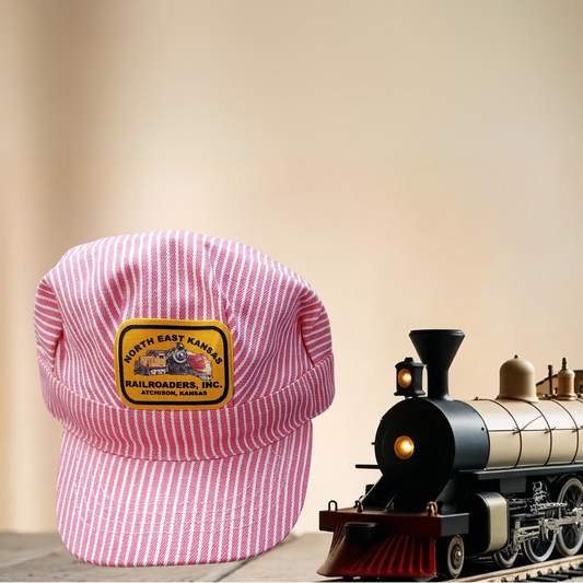 Pink and white striped cap with a gold and navy North East Kansas Railroaders patch next to a model train on a wooden surface.