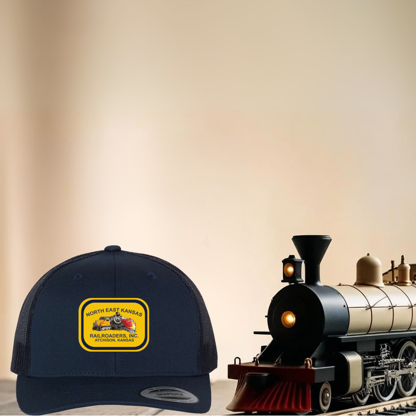 Navy blue cap with gold and navy North East Kansas Railroaders logo next to a toy train on a beige background