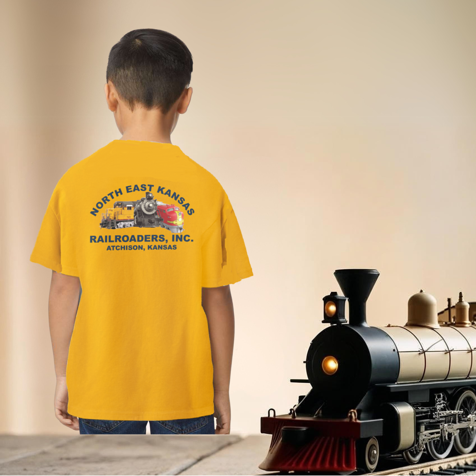 Child wearing a yellow t-shirt with a train design, standing next to a model train on a table.